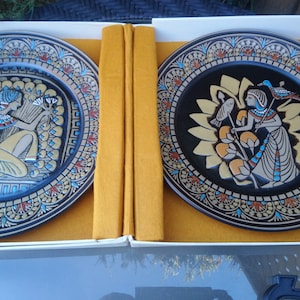 May include: Two decorative ceramic plates with black backgrounds and gold and blue accents. Each plate features a stylized image of a woman in an Egyptian-inspired costume.