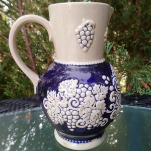 Puede incluir: Una jarra de cer&aacute;mica blanca y azul con asa y dise&ntilde;o de uvas. La jarra est&aacute; decorada con uvas y hojas blancas sobre un fondo azul.