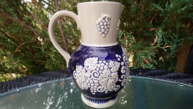 Puede incluir: Una jarra de cer&aacute;mica blanca y azul con asa y dise&ntilde;o de uvas. La jarra est&aacute; decorada con uvas y hojas blancas sobre un fondo azul.