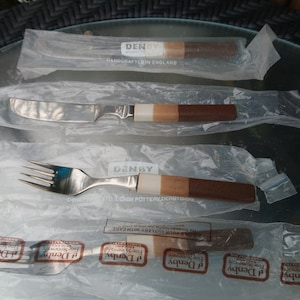 May include: Three sets of Denby cutlery in individual plastic packaging. The cutlery includes a knife, fork, and spoon. The handles are made of wood and have a brown and white striped pattern. The packaging has the Denby logo and the text "Handcrafted in England".