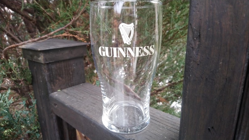 Puede incluir: Un vaso de pinta de vidrio transparente con el logotipo de Guinness y un dise&ntilde;o de arpa. El vaso est&aacute; vac&iacute;o y se encuentra sobre una superficie de madera.