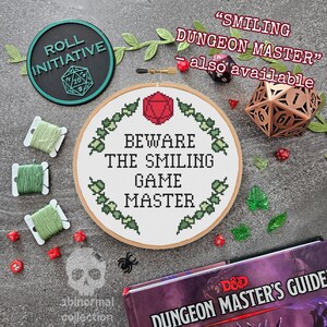 Op de afbeelding: Een borduurring met kruissteek met een witte achtergrond en groene borduurwerk dat een krans van bladeren rond de tekst "Beware the Smiling Game Master" afbeeldt. De ring ligt op een grijs oppervlak met verschillende dobbelstenen, garen en andere knutselmaterialen.