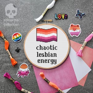 May include: A cross-stitch embroidery hoop with a lesbian pride flag design and the text "chaotic lesbian energy." The image includes embroidery floss in various colors, stickers, and a small "LOVE" pin.