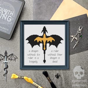 May include: A framed cross-stitch piece featuring a black and gold dragon silhouette centered above the text: a dragon without its rider is a tragedy, a rider without their dragon is dead. The design is displayed in a dark blue square frame.