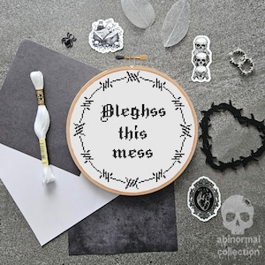 以下が含まれることがあります： 黒い糸で刺繍された白い刺繍枠で、ゴシック体で「Blehgss this mess」という文字が書かれています。枠は黒い有刺鉄線とその他のゴシック調の装飾で囲まれています。
