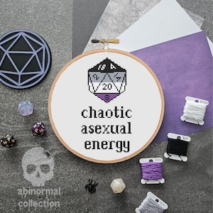 May include: A white embroidery hoop with a black and white 20-sided die with a purple and gray acexual pride flag design. The text "chaotic asexual energy" is embroidered below the die.