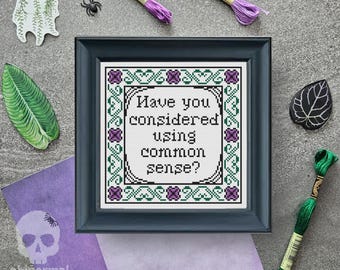 May include: A black framed cross-stitch embroidery with a white background and green and purple accents. The embroidery reads "Have you considered using common sense?"