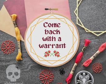 May include: A white embroidery hoop with a gold geometric frame and the text "Come back with a warrant" in red. The hoop is surrounded by red and yellow embroidery floss.