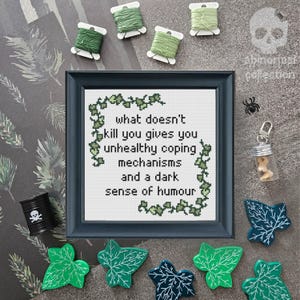 May include: A framed cross-stitch with a dark green ivy border and the text "what doesn't kill you gives you unhealthy coping mechanisms and a dark sense of humour".