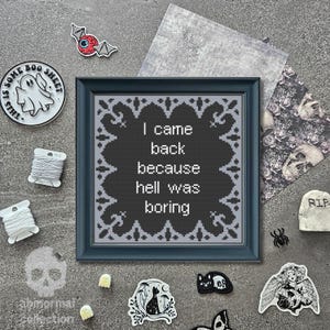 May include: A framed cross-stitch piece with the text "I came back because hell was boring." The dark gray framed artwork is surrounded by Halloween-themed stickers, a ghost pin, and spools of thread. A small tombstone and a spider are also present.