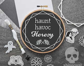 May include: Black and white cross-stitch embroidery hoop with the text "Haunt Havoc Heresy" and two small floral designs. The hoop is on a gray surface with other black and white decorative items.