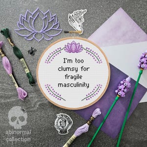 以下が含まれることがあります： 紫と白の刺繍枠に「I'm too clumsy for fragile masculinity」という文字が書かれており、花の模様で囲まれています。枠は灰色の表面に置かれており、紫と白の刺繍糸、紫のグラデーションの白い紙、紫の花があります。