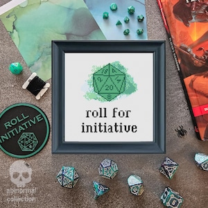 Op de afbeelding: Een groene 20-zijdige dobbelsteen met een witte omtrek en zwarte cijfers is geborduurd op een wit canvas met de tekst "roll for initiative" in zwart. Het canvas is ingelijst in een donkerhouten lijst.