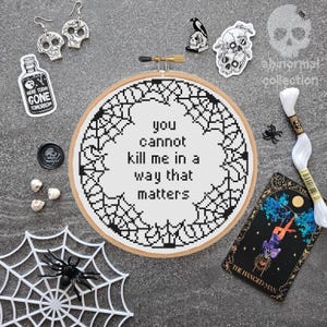 Könnte beinhalten: Ein Kreuzstich-Stickrahmen mit dem Text "you cannot kill me in a way that matters", umgeben von einem Spinnennetz-Design. Das Bild enthält Schädelohrringe, Aufkleber, eine Tarotkarte und weiteres Gothic-Zubehör.