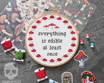 May include: A white embroidery hoop with red mushrooms around the edge and the text "everything is edible at least once" in the center. The hoop is surrounded by embroidery floss, mushroom stickers, and a mushroom-themed glass dome.