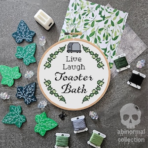 Puede incluir: Un aro de bordado de punto de cruz con el texto "Live Laugh Toaster Bath" rodeado de hojas verdes. El aro está sobre una superficie gris con carretes de hilo de bordar verde y negro, hojas verdes y una pequeña tostadora blanca.