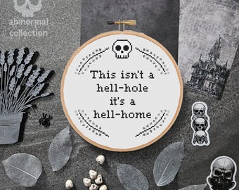Puede incluir: Un aro de bordado blanco con texto negro que dice "This isn't a hell-hole it's a hell-home". El aro está rodeado de hojas y cráneos negros y blancos.