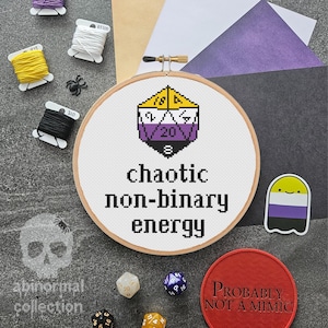 May include: A white embroidery hoop with a cross-stitch design of a purple, yellow, and black dice with the text "chaotic non-binary energy".