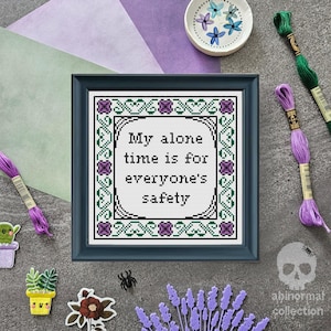 May include: A cross-stitch pattern with a blue frame and a white background. The pattern features a green and purple floral border and the text "My alone time is for everyone's safety".