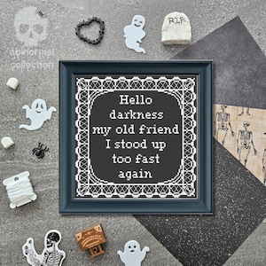 May include: A framed cross-stitch piece featuring a black background with white text that reads: Hello darkness my old friend I stood up too fast again. The embroidery is enclosed in a dark frame with a decorative white border pattern.