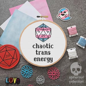 May include: A white embroidery hoop with a cross-stitch design of a 20-sided die with the colors of the transgender pride flag. The text "chaotic trans energy" is stitched below the die.