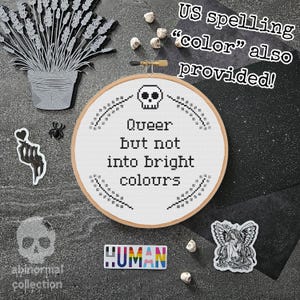 Könnte beinhalten: Ein weißer Stickrahmen mit dem Text "Queer but not into bright colours" in Schwarz. Der Rahmen ist umgeben von schwarzen und weißen Totenköpfen, einer schwarzen Spinne und einem schwarz-weißen Aufkleber einer Hand, die ein Herz hält. Der Text "US spelling "color" also provided!" ist in Weiß auf schwarzem Hintergrund geschrieben.