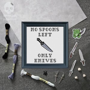May include: A blue framed cross-stitch with the text "No Spoons Left Only Knives" and a black and white pixelated image of a knife. The cross-stitch is surrounded by various craft supplies, including embroidery floss, a frog charm, and small knives.