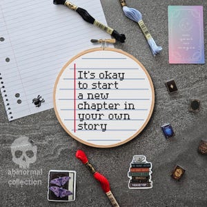 Puede incluir: Un aro de punto de cruz con el texto "It's okay to start a new chapter in your own story." La imagen incluye hilo de bordar, pegatinas y libros en miniatura. La paleta de colores incluye blanco, negro, rojo y azul.