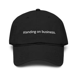 May include: Black baseball cap with the phrase "standing on business" in white, centered on the front. The cap has a curved brim and a button on top. A casual accessory.