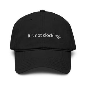 May include: Black baseball cap with a curved brim. The cap features the white text, "it's not clocking." The hat is made of a soft material and has a structured crown. A casual accessory for everyday wear.