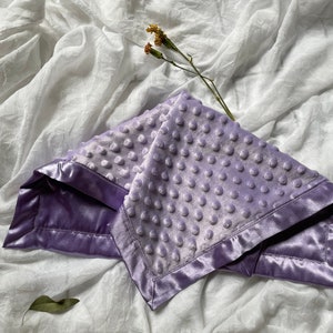 May include: A lavender baby blanket with a textured, dotted surface and a satin border. The blanket is folded and rests on a white, wrinkled fabric. A dried flower stem is placed on the blanket.