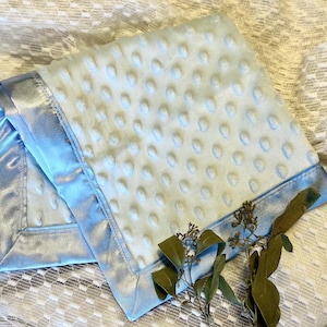 Pode incluir: Um cobertor azul claro para bebé com uma superfície texturizada com pontos e uma borda de cetim. O cobertor está dobrado, mostrando a borda de cetim e o material macio e felpudo. O cobertor tem cerca de 76 cm quadrados.