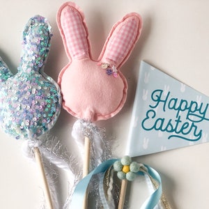 May include: Three Easter bunny wands with blue and pink fabric and sequins. The wands have wooden sticks and white and blue ribbons. A blue and white pennant with the text "Happy Easter" is in the background.