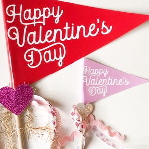 May include: Two Valentine's Day pennants, one red and one pink, with the text "Happy Valentine's Day" in white cursive font. Each pennant is attached to a wooden stick with pink and white ribbon and a sparkly pink heart.