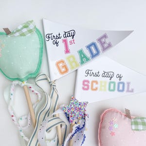 Op de afbeelding: Twee vlaggen in de vorm van een pennant met regenboogletters. De eerste vlag zegt "First day of 1st GRADE" en de tweede vlag zegt "First day of SCHOOL".