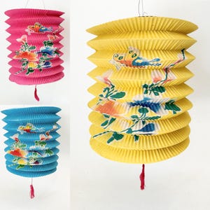 May include: Three cylindrical paper lanterns in pink, blue, and yellow, each featuring a pleated accordion design. The lanterns are decorated with colorful printed illustrations of butterflies and floral branches, and each includes a small red tassel.