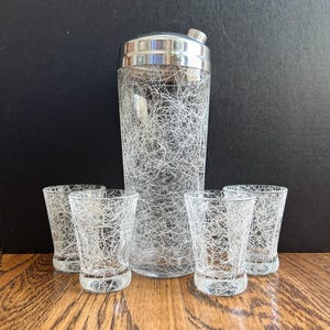 May include: A clear glass cocktail shaker with a silver metal lid and four matching drinking glasses. Each piece features a white, thin, chaotic, web-like or stringy pattern overlaying the clear glass surface, creating a cohesive set.