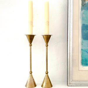 May include: Two vintage brass candlesticks with cream-colored candles. The candlesticks have a hammered texture on the base and a slender, textured stem. The candles are unscented and have a decorative pattern near the top.