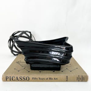 May include: A glossy black ceramic bowl with a unique, layered design. The bowl sits on a vintage book titled "Picasso Fifty Years of His Art". A decorative black and white sphere is in the background.