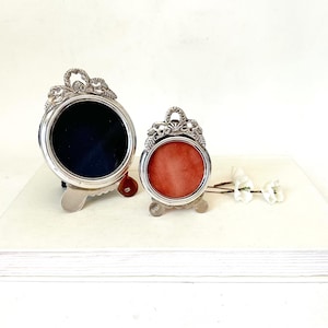 May include: Two silver-toned metal picture frames with ornate, leaf-like detailing at the top. The larger frame features a dark navy blue circular insert, while the smaller frame contains a warm, burnt orange circular insert. Both stand upright.