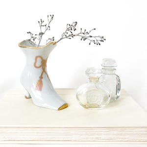 May include: A white porcelain boot-shaped vase with gold trim and a pink and gold bow design, holding silver-grey branches. Two clear glass perfume bottles sit beside the vase, creating a decorative display.