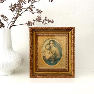 May include: A rectangular gold-toned frame with ornate carved edges containing an oval print of a person holding a child. The print features soft muted tones and is set against a textured gold background with embossed floral and geometric patterns.