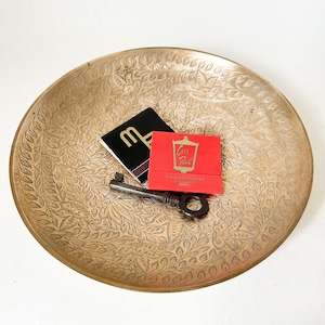May include: A circular brass tray with an embossed floral design. A black and white notepad with a stylized "M" is next to a red matchbook with gold lettering. A vintage key lies on the tray.