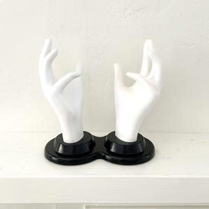 May include: A decorative sculpture featuring two white ceramic hands in a gesture of offering or receiving. The hands are mounted on a black, oval-shaped base. The sculpture is set against a white background.
