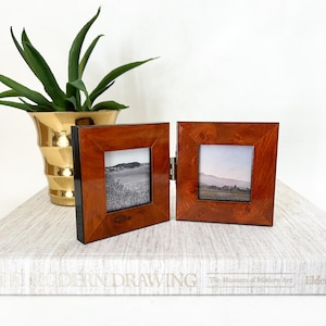 May include: A hinged, double-sided wooden photo frame with a glossy finish. The frame is a warm brown color and holds two square black and white landscape photos. A gold-colored vase with a green plant is in the background.