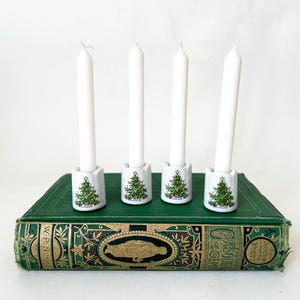 May include: Four white taper candles stand in individual white ceramic holders, each decorated with a small green Christmas tree illustration. These holders are arranged in a row on top of a thick, green, antique-style hardcover book.