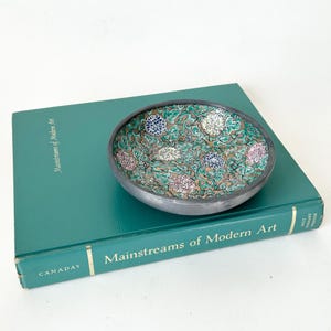 May include: A shallow, round ceramic bowl with a dark gray exterior and a detailed interior pattern featuring green swirling vines and small, multi-colored floral clusters in shades of blue, white, and pink against a tan background.