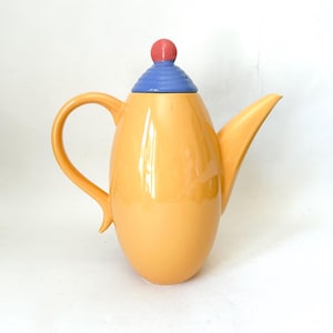 May include: A glossy yellow ceramic teapot with a curved handle and a pointed spout. The lid features a blue, ribbed, conical base topped with a small, round, coral-colored knob. The body of the teapot has a smooth, rounded, vertical shape.