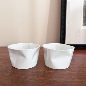 May include: Two white ceramic bowls with a distinctively irregular, crumpled texture. Each bowl features a smooth, glossy finish and a wide, open top with soft, undulating edges that mimic the appearance of crushed paper or soft material.