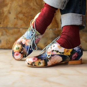 Può includere: Un paio di scarpe con lacci a stampa floreale con calze rosse. Le scarpe hanno una base bianca con una varietà di fiori e foglie colorati stampati su di esse. I lacci sono di colore blu chiaro.